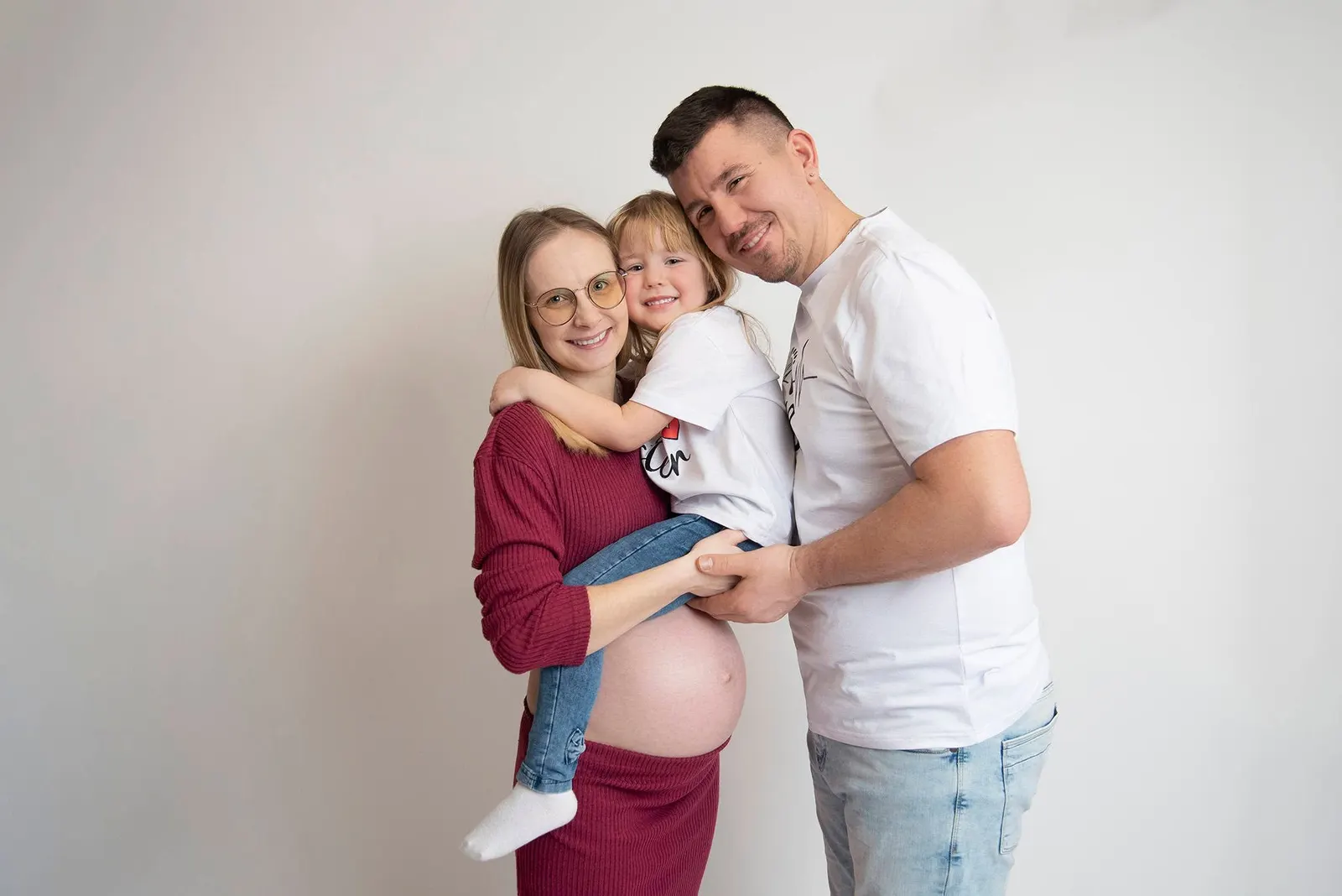 Mama w ciąży, tata i córeczka przytuleni na jasnym tle w studiu